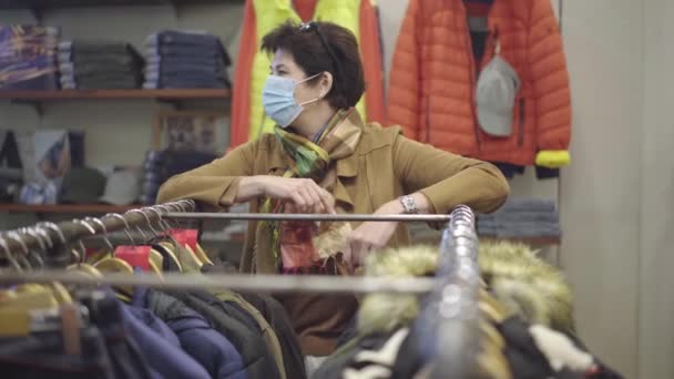 Retrato de mulher bonita meio adulto em Covid-19 máscara facial em pé na loja de roupas no rack com roupas penduradas. Cliente caucasiano feminino confiante comprando roupa em vendas coronavírus Black Friday. — Vídeo de Stock