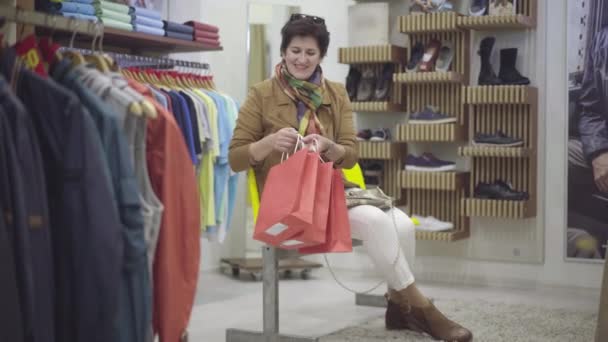 Amplio tiro de feliz caucásica hembra comprador admirando las compras en la tienda de ropa. Retrato de la alegre mujer caucásica sentada en el centro comercial con bolsas de compras y sonriendo a la cámara. Felicidad y estilo de vida. — Vídeo de stock