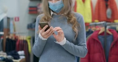 Coronavirus yüz maskesi takmış beyaz bir kadın giyim mağazasında akıllı telefon kullanıyor. Covid-19 Kara Cuma 'sında internetten alışveriş yapan bir kadın. Sinema 4k ProRes Merkezi.