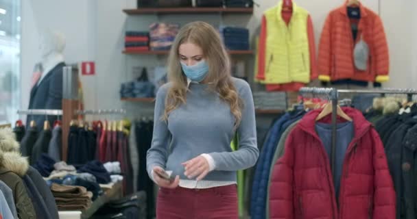 Covid-19 의젊은 캅카스 여성은 옷 가게에서 스마트폰을 사용하여 마스크를 쓴다. 온라인에서 긍정적 이고 아름다운 여성 손님이 문자를 보내는 모습이 블랙 프라이데이 쇼핑으로 등장 한다. 시네마 4k ProRes HQ. — 비디오