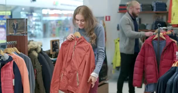 옷 가게에서 코트를 보고 있는 젊은 여성을 유혹하고 카메라로 웃는다. 의상 가게에서 남자가 배경 의상을 고르는 자신있는 부자 고객의 모습. 시네마 4k ProRes HQ. — 비디오
