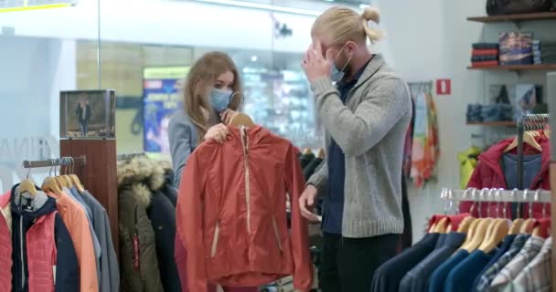 Pasangan muda Kaukasia berbelanja di toko pakaian selama penjualan Jumat Hitam pada pandemi coronavirus. Pria dan wanita di Covid-19 masker wajah memilih mantel di pusat perbelanjaan. Sinema 4k ProRes HQ. — Stok Video