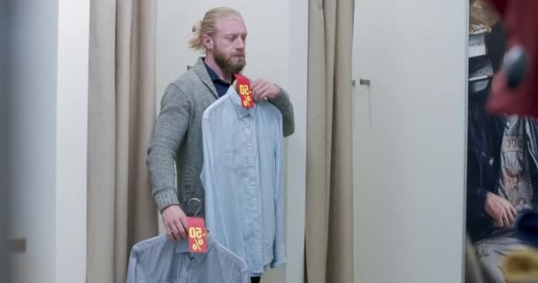 Joven hombre confiado eligiendo camisetas durante las ventas del Viernes Negro. Retrato de comprador varón caucásico confiado entrando en vestidor de la tienda de ropa y cortina de cierre. Sede del cine 4k ProRes. — Vídeo de stock