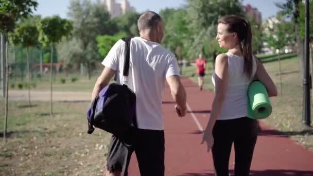 Bakåt syn på positiva kaukasiska man och kvinna promenader på soliga idrottsplanen och prata. Kamera följer positiv idrottsman och idrottskvinna promenader efter träning utomhus. — Stockvideo