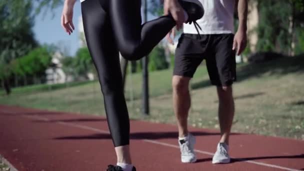 Onherkenbare slanke vrouw in zwarte legging die zich uitstrekt over zonnige hardloopbaan met een man die opwarmt op de achtergrond. Sportief Kaukasisch echtpaar dat voor de training buiten traint. — Stockvideo