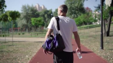 Spor çantası ve su şişesiyle koşu yolunda yürüyen kendine güvenen bir sporcunun arka görüntüsü. Spor kıyafetleri içinde beyaz bir adam antrenmandan sonra gezintiye çıkıyor ya da güneşli bir günde dışarıda antrenman yapıyor..