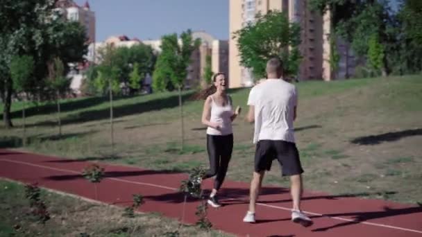 Breed shot van vrolijke atletische fit man en vrouw joggen buiten op de zomerochtend. Gelukkig Kaukasisch echtpaar trainen en trainen op zonnige dag op hardloopbaan op sportveld. — Stockvideo