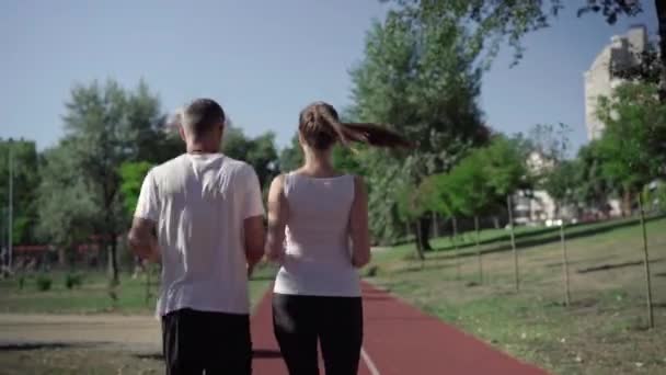 Achteraanzicht van een volwassen stel joggers die op de zomerochtend langs de baan rennen. Camera volgt fitte atletische blanke man en vrouw genieten van training buiten. Actieve levensstijl. — Stockvideo