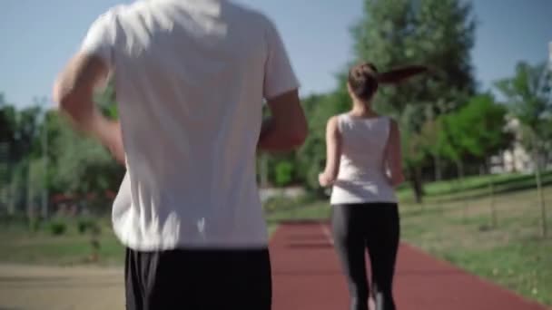 Widok z tyłu nierozpoznawalnego sportowca biegnącego za szczupłą białą kobietą na słonecznym torze. Pozytywna sportowiec zawraca i macha do przyjaciela. Radość i koncepcja treningu. — Wideo stockowe