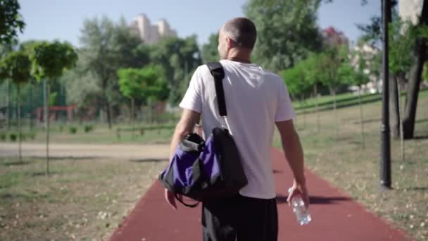 Achteraanzicht van zelfverzekerde volwassen sporter wandelen met sporttas en waterfles langs hardloopbaan. Blanke man in sportkleding wandelen na training of training buiten op zonnige dag. — Stockvideo