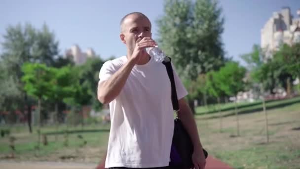 Portrait of handsome confident Caucasian sportsman walking on running track, drinking refreshing water, and leaving with sport bag. Adult man strolling after workout outdoors. — Stockvideo