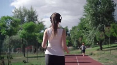Kamera, yaz sabahı koşu pistinde koşan, kulaklıklı, zayıf, mutlu, beyaz kadın sporcuyu takip ediyor. Sağlıklı bir kadının dışarıda egzersiz yapması. Egzersiz konsepti.