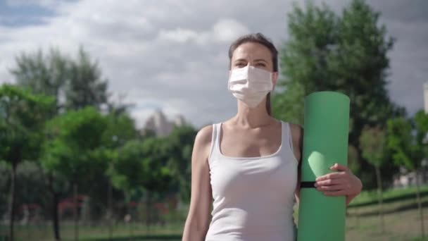 Femme sportive caucasienne confiante marchant avec tapis d'exercice à l'extérieur. décoller le masque facial covid-19 et respirer. charmante femme adulte regardant la caméra et souriant après l'entraînement coronavirus. — Video