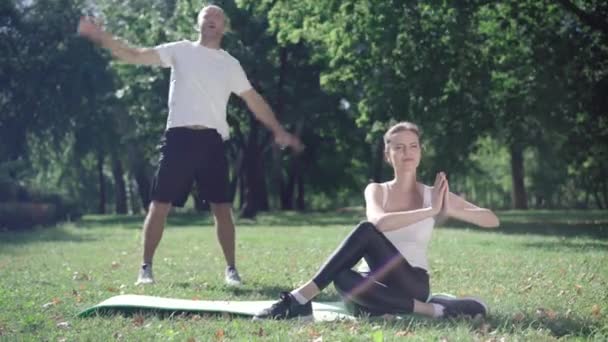 Szerokie ujęcie pozytywnej sportowca siedzącego na pozycji jogi jako silny sportowiec rozciągający się w tle. Szczęśliwa para kaukaska trenuje na świeżym powietrzu w słoneczny letni dzień. Koncepcja sportu. — Wideo stockowe