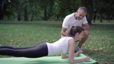 Pozitif kişisel antrenör, sporcuyu dışarıda egzersiz minderinin üzerinde kalas pozisyonunda tutuyor. Gülümseyen yakışıklı sporcu sporcu kadına antrenmanda yardım ediyor. Spor konsepti.