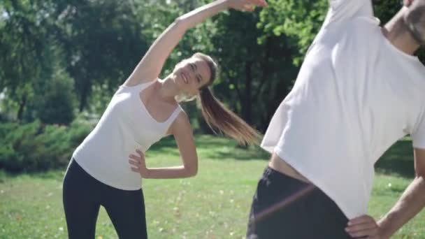 Smiling Caucasian sportswoman repeating warmup exercise after blurred man bending at front as having sudden sharp pain in stomach. Unwell woman overworking during morning training. — Stock Video