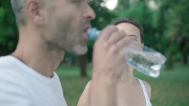 Piękna uśmiechnięta kobieta dopasowująca kucyk jako zamazanego mężczyznę pijącego orzeźwiającą wodę z przodu. Portret kobiety rasy białej po treningu ze sportowcem na świeżym powietrzu. — Wideo stockowe