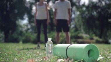 Arka planda bulanık beyaz bir çift uzanırken güneş ışığında uzanan su şişesi ve egzersiz paspası yakın plan. Açık havada güneşli bir yaz sabahı ısınan zinde bir erkek ve kadın..