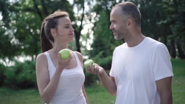 トレーニング後にグリーンリンゴを食べカメラで微笑んだ幸せなスポーツマンと女性。屋外でのワークアウト後の積極的な白人スポーツマンとスポーツウーマンの肖像. — ストック動画