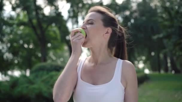 Portret van een prachtige vrolijke sportvrouw die gezonde groene appel eet en lacht naar de camera. Gelukkig ontspannen vertrouwen Kaukasische vrouw poseren na training of training buiten. — Stockvideo