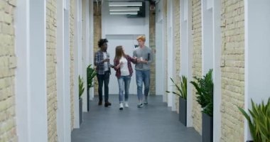 Modern ofiste koridorda yürüyen ve sohbet eden üç pozitif genç. Kafkasyalı ve Afrikalı Amerikalı erkek ve kadınlar tatilden işe dönerken konuşuyor ve gülüyorlar. Sinema 4k ProRes Merkezi.