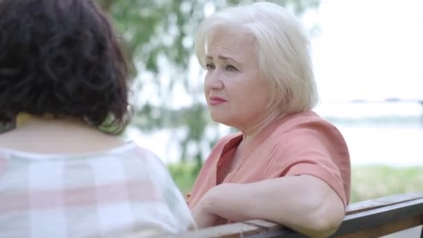 Внимательная заботливая пожилая женщина слушает неузнаваемую девушку и качает головой. Портрет кавказского пенсионера, делящегося секретами и проблемами с другом в солнечном летнем парке. — стоковое видео
