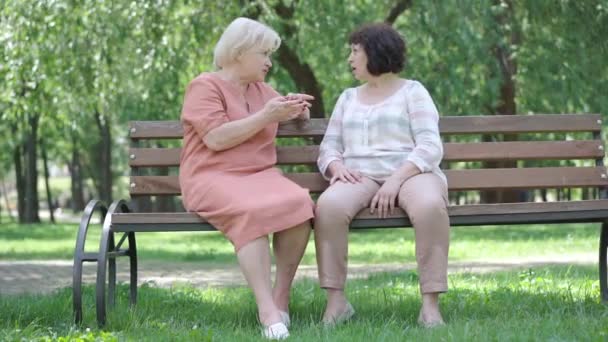 Широкий постріл блондинки старша жінка кричить на вухо глухому другу. Портрет розслаблених кавказьких пенсіонерів, що сиділи на лавці в сонячному парку і розмовляли. Старіння і дружба. — стокове відео