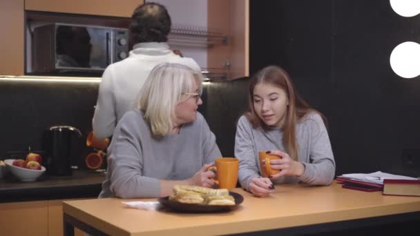 Madre positiva bebiendo té con hija adolescente en la cocina como padre tomando taza de casillero en el fondo. Familia caucásica disfrutando de la noche tranquila en casa. Concepto de estilo de vida. — Vídeos de Stock