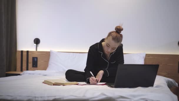 Ampla foto de bela menina caucasiana confiante sentada na cama com laptop e livros. Retrato de adolescente magro em pijama fazendo lição de casa no quarto em casa. Conceito de educação e aprendizagem online. — Vídeo de Stock