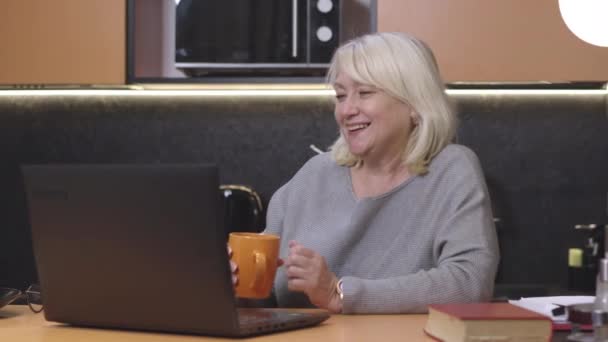 Femme âgée blonde positive utilisant le chat vidéo comme boire du thé dans la cuisine à la maison. Portrait de dame blanche joyeuse appréciant la communication sans fil et souriant. Mode de vie et technologies modernes. — Video