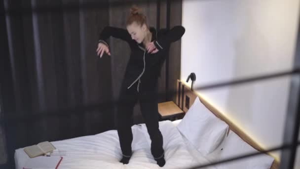 Радісний танець безтурботної Кавказької дівчини - підлітка на ліжку. Широкий постріл веселих струнких підлітків, що танцюють у спальні. Розслабся, коли підліток розважається вдома. Життєпис і концепція підліткового віку. — стокове відео
