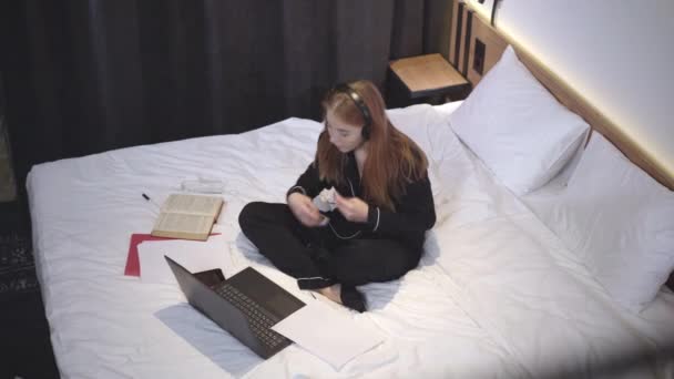 Najlepszy widok beztroskiej, białej nastolatki siedzącej na łóżku w słuchawkach, słuchającej muzyki z laptopa i robiącej selfie na smartfonie Szerokie ujęcie radosnej nastolatki cieszącej się wieczorem w domu. — Wideo stockowe