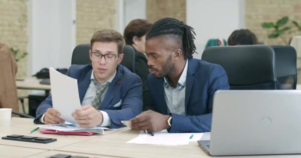 Jonge Kaukasische en Afro-Amerikaanse mannen zitten in functie en bespreken het opstarten. Zelfverzekerde succesvolle zakenmensen die praten en projecten analyseren met collega 's op de achtergrond. Cinema 4k ProRes Hoofdkwartier. — Stockvideo