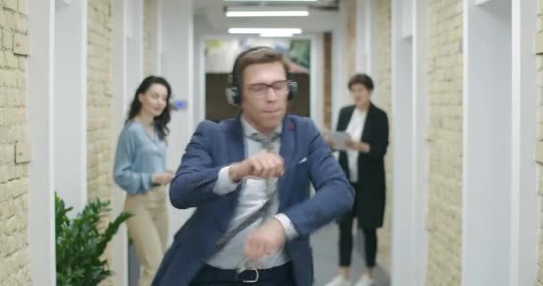 Χαρούμενος όμορφος άντρας με ακουστικά που χορεύει στο διάδρομο του γραφείου. Χαρούμενοι πολυεθνικοί συνάδελφοι που ενώνονται με το χορό και πετάνε έγγραφα. Ομάδα θετικών συναδέλφων γιορτάζουν την επιτυχία. Κινηματογράφος 4k ProRes HQ — Αρχείο Βίντεο