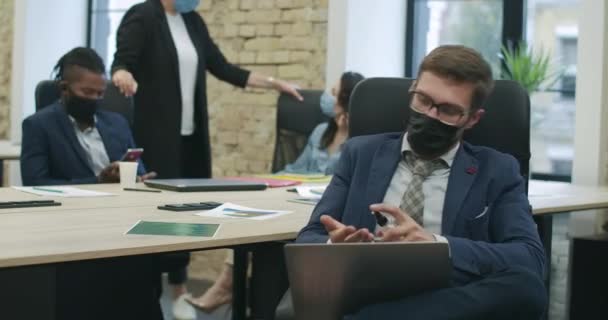 Homme confiant dans les lunettes et le masque facial Covid-19 désinfectant les mains avec un désinfectant et tapant sur le clavier de l'ordinateur portable. Mes collègues discutent des affaires en arrière-plan. Style de vie agréable. Siège social Cinema 4k ProRes. — Video