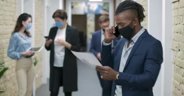 Magabiztos afro-amerikai üzletember Covid-19-es maszkban beszél telefonon az irodai folyosón, kollégákkal a háttérben. Coronavirus üzleti életmód. Cinema 4k ProRes főhadiszállás. — Stock videók