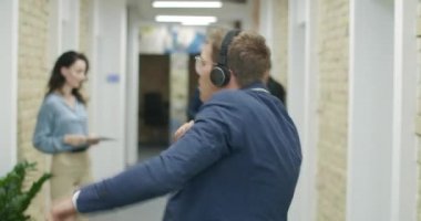 Kulaklıkla dans eden neşeli genç iş adamı arka plandaki cihazlarla video kaydediyor. İş arkadaşları ofis koridorunda mutlu çalışanları filme alıyor. Sinema 4k ProRes Merkezi.