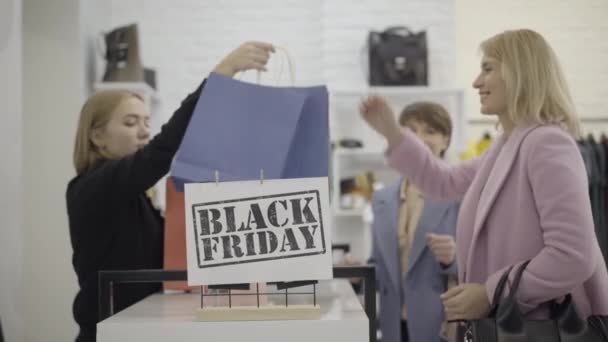 Gülümseyen tezgahtar mağazadaki mutlu müşterilere alışveriş torbaları uzatıyor. Bulanık beyaz kadınlar alışveriş merkezindeki Kara Cuma satışları sırasında kıyafet alıyorlar. Moda ve ticaret konsepti. — Stok video