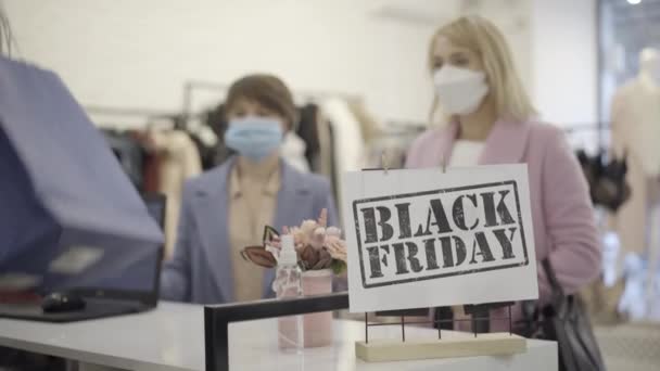 Fekete Péntek banner a pénztárgépen, homályos boldog nőkkel Covid-19-es maszkban, akik bevásárlószatyrokat vesznek el a bolti asszisztenstől. Elégedett kaukázusi vásárlók ruhákat vásárolnak a koronavírus világjárvány idején. — Stock videók