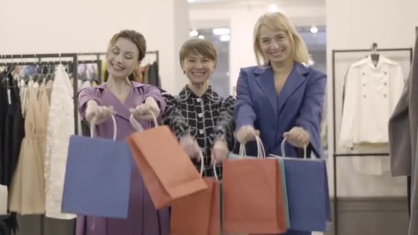 Tiga wanita Kaukasia positif dengan pakaian elegan Berpose dengan tas belanja di mal. Potret bahagia percaya diri indah pembeli membeli pakaian selama Black Friday penjualan. — Stok Video