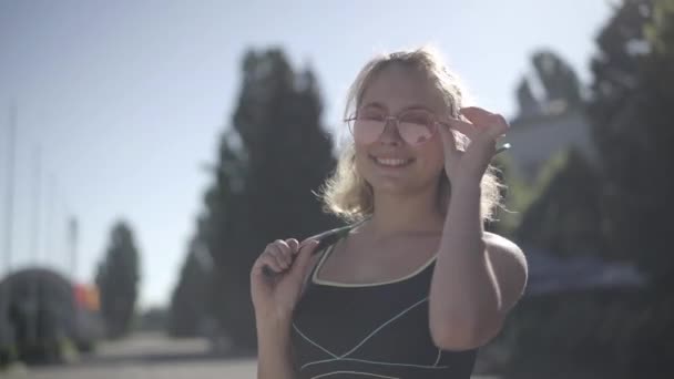 Збільшений портрет щасливої красивої молодої жінки в сонцезахисних окулярах з тенісною ракеткою на сонці. Впевнена, що кавказька спортсменка стоїть надворі, дивиться на камеру і посміхається. — стокове відео