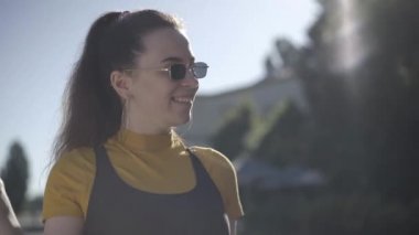 Güneş gözlüklü, açık havada poz veren yakışıklı, beyaz tenli genç bir kadının yakın çekimi. Lolipop emen ve kameraya gülümseyen sıska esmer kadının portresi. Flört ve yaşam tarzı.