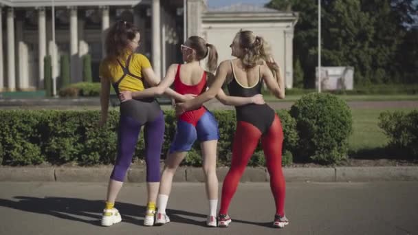 Güneşli şehir caddesinde dans eden ve kameraya gülümseyen üç beyaz kadının geniş açılı görüntüsü. Spor giyim sektöründeki kaygısız arkadaşlarınız dışarıda eğleniyor.. — Stok video