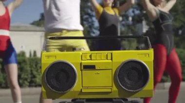 Sarı retro teyp, arka planda disko müzik egzersizleri yapan bir grup formda insanla gün ışığında duruyor. 1980 'lerde açık havada neşeli erkek ve kadınların eğitimi. Eski konsept.