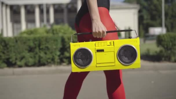Eine nicht wiederzuerkennende schlanke Frau in roten Leggings läuft mit einem gelben Tonbandgerät über die sonnige Sommerstraße. Fit kaukasische 80er-Jahre-Sportlerin flaniert und tanzt zu Musik im Freien. — Stockvideo