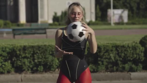 Mellanslag av stark säker idrottskvinna poserar med fotboll på solig sommargata. Porträtt av vacker smal vit kvinna i solglasögon och sportkläder som håller fotboll och ler. — Stockvideo