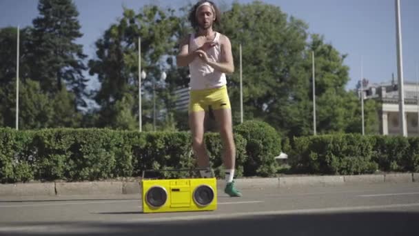 Wide shot of active training of fit Caucasian sportsman to disco music in 1980s. Portrait of handsome confident man exercising on urban street with retro yellow tape recorder standing on road. — Stock Video