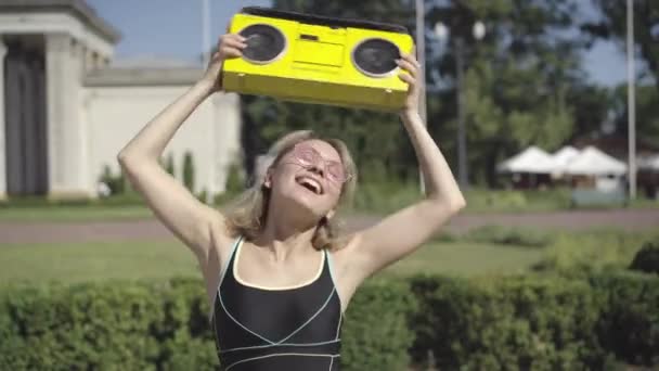 Carefree happy 1980s woman dancing slowly and signing to disco music holding retro yellow tape recorder over head. Portrait of relaxed Caucasian lady enjoying sunny summer day outdoors. — Stock Video