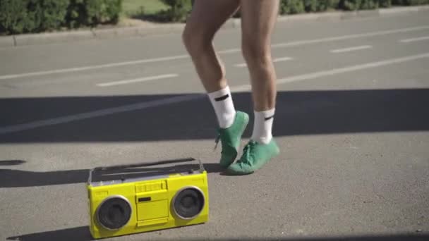 Piernas de ajuste irreconocible joven caucásico haciendo ejercicios deportivos en el soleado día de verano al aire libre con grabadora de cinta amarilla vintage de pie en la carretera. Estilo de 1980 deporte y estilo de vida. — Vídeos de Stock