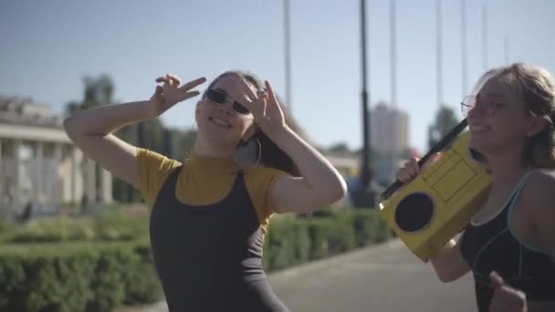 Дві жінки в сонцезахисних окулярах 1980-х танцюють, щоб ретро-диско музика з вінтажного жовтого магнітофона. Портрет радісних молодих кавказьких друзів, які бавляться на міському вулиці сонячним світлом.. — стокове відео